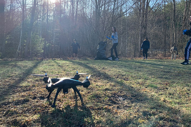 Drone on the grass while students collect trash among the trees